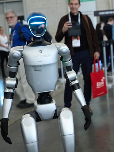 A Unitree humanoid walking a trade-show aisle while a visitor films it
